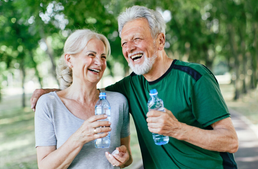 A senior woman and a senior man smiling and walking in a park while holding water bottles.