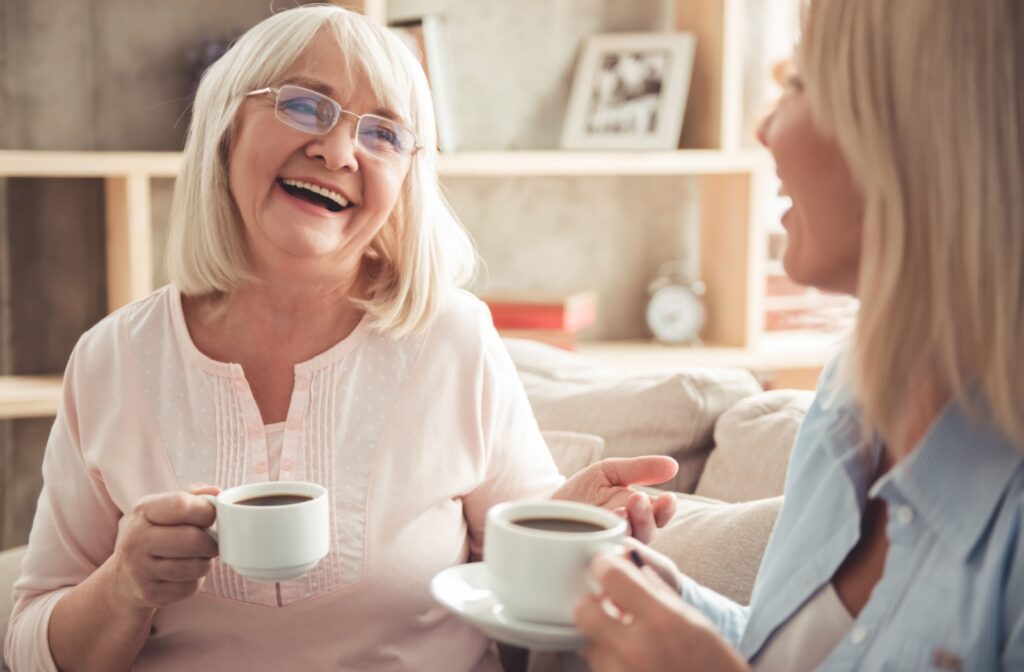 An older adult woman and her daughter sitting on a couch smiling and talking to each other while holding a cup of coffee.