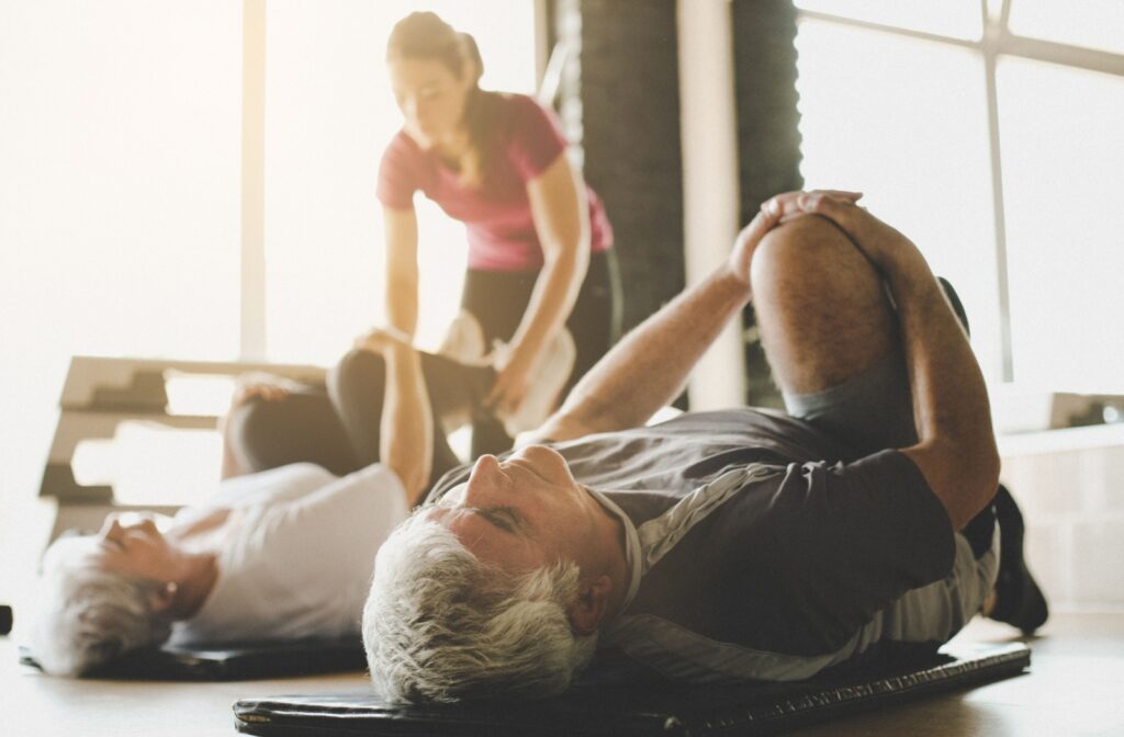 2 seniors lay on their backs in a well-lit yoga studio, stretching their legs to their chests.